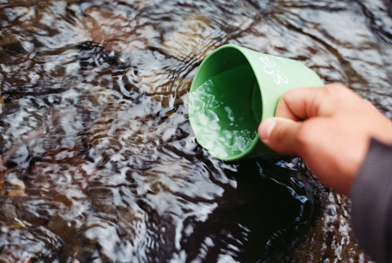 A hand scooping water from a flowing stream into a cup outdoors